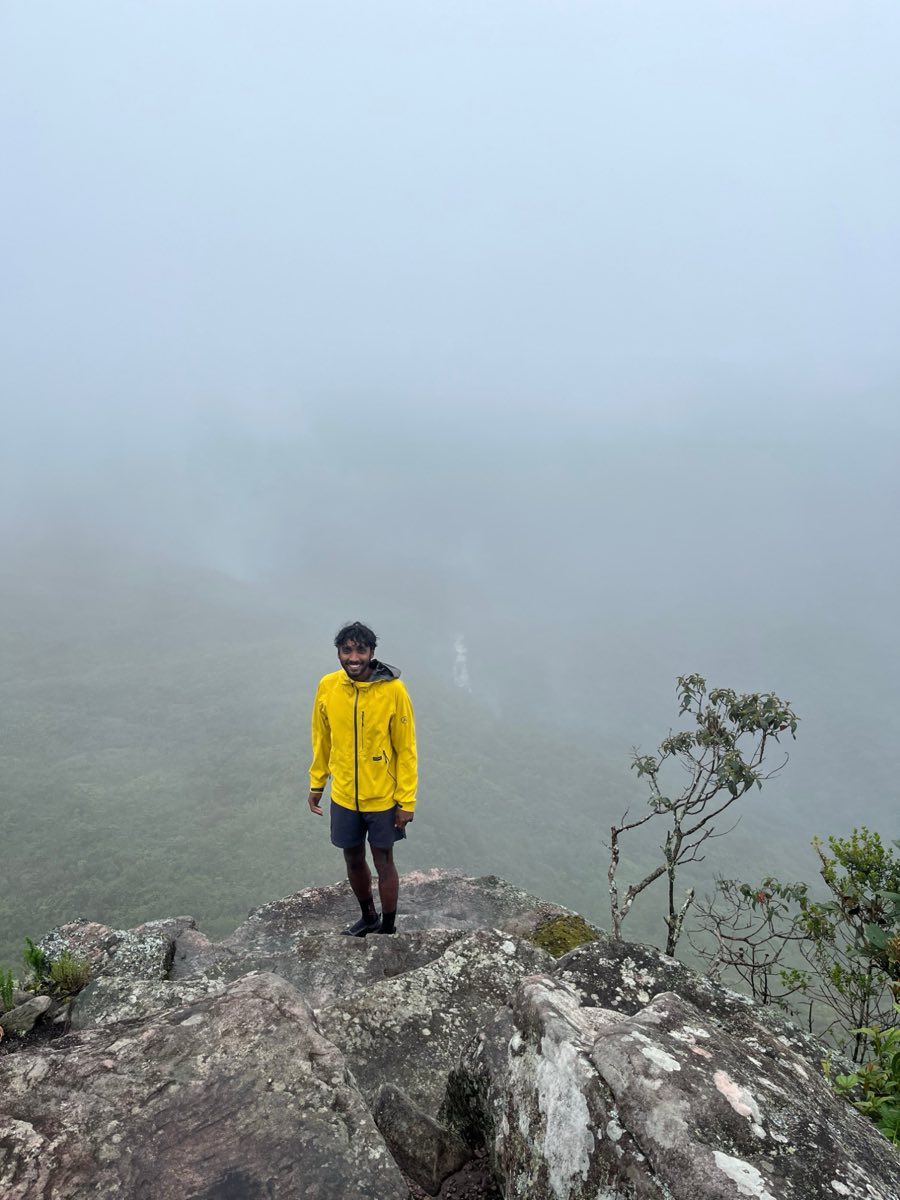 doglike standing on a wet rock at the edge of a ridge in a yellow rain jacket and shorts, smiling, with fog closing over the green valley behind him.